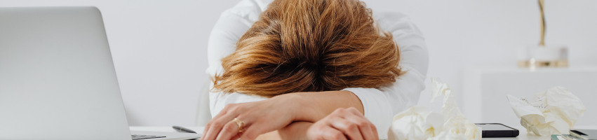 Frau die mit Armen und Kopf auf dem Schreibtisch liegt und erschöpft nicht weiter arbeiten kann